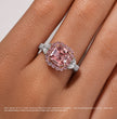 A close-up of a ring worn on a finger, featuring a large rectangular pink gemstone surrounded by a halo of small pink and clear stones, with additional small side stones on a silver-toned band. A note at the bottom explains the size and color reference of the center stone.