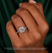 A close-up of a hand wearing a silver halo ring with a cushion-cut diamond center stone, set against a dark green satin background with a small disclaimer note at the bottom.