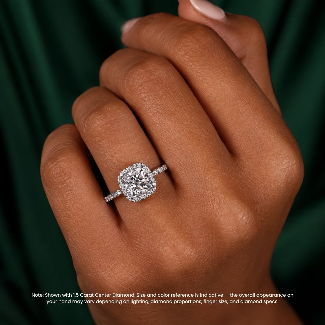A close-up of a hand wearing a silver halo ring with a cushion-cut diamond center stone, set against a dark green satin background with a small disclaimer note at the bottom.