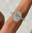 A close-up of a diamond ring with a large oval center stone and halo setting, worn on a finger against a soft light gray background.