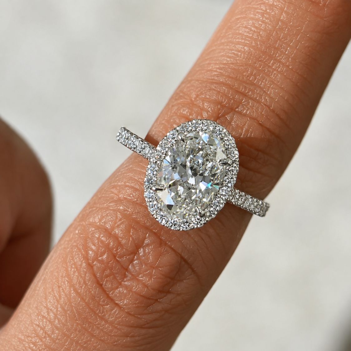 A close-up of a diamond ring with a large oval center stone and halo setting, worn on a finger against a soft light gray background.