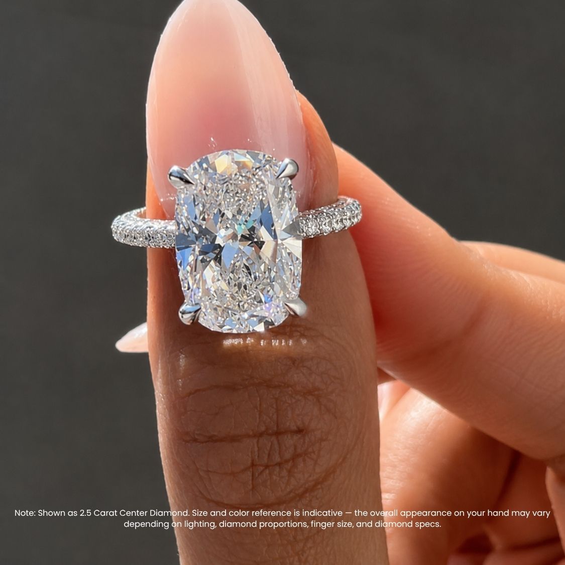 An engagement ring with a large cushion-cut diamond and a silver-toned pavé band, shown worn on a finger against a dark gray background. A note at the bottom explains the carat size reference.