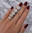 A hand with dark burgundy nail polish displays five silver rings with round-cut diamonds of varying carat sizes (1ct to 5ct) arranged vertically. The background is a white and gray marble surface.
