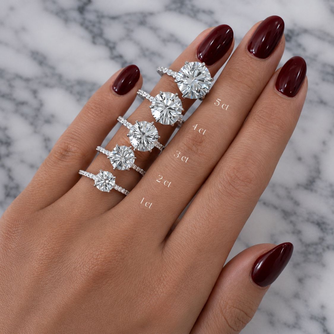 A hand with dark burgundy nail polish displays five silver rings with round-cut diamonds of varying carat sizes (1ct to 5ct) arranged vertically. The background is a white and gray marble surface.